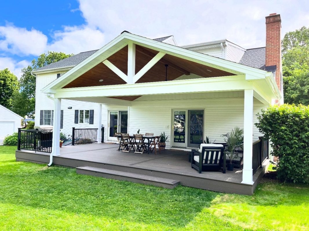 White house with a large covered wood deck; outdoor dining table, chairs, and lounge seating, green lawn and blue sky nearby.
