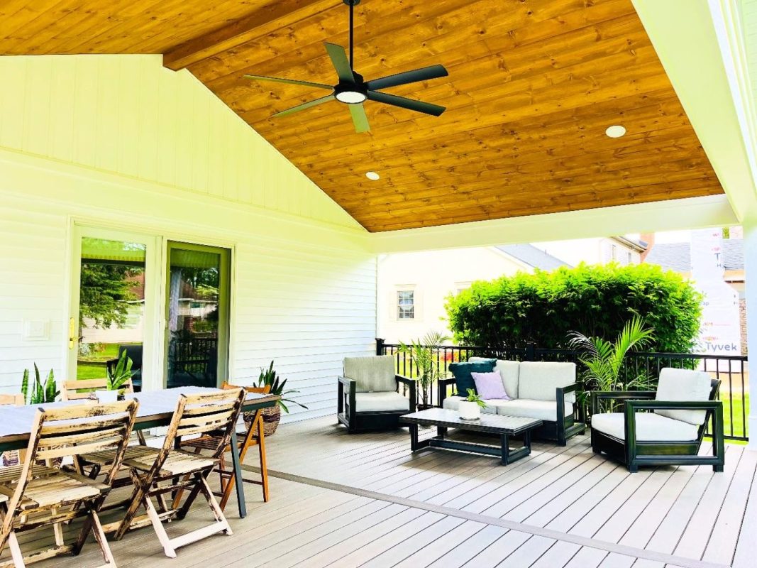 Covered outdoor patio with a wooden ceiling, ceiling fan, and white walls featuring a long dining table and rustic chairs on the left; a lounge seating area on the right with cushions and plants.