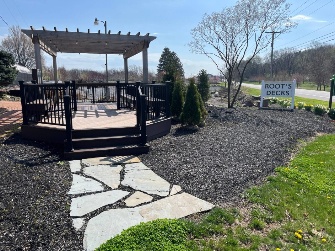 Raised wooden deck with a pergola and black railings, in a landscaped yard near a 'Root's Decks' sign.