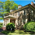 A stone manor with white columns and a covered porch, surrounded by trimmed bushes under a bright blue sky.
