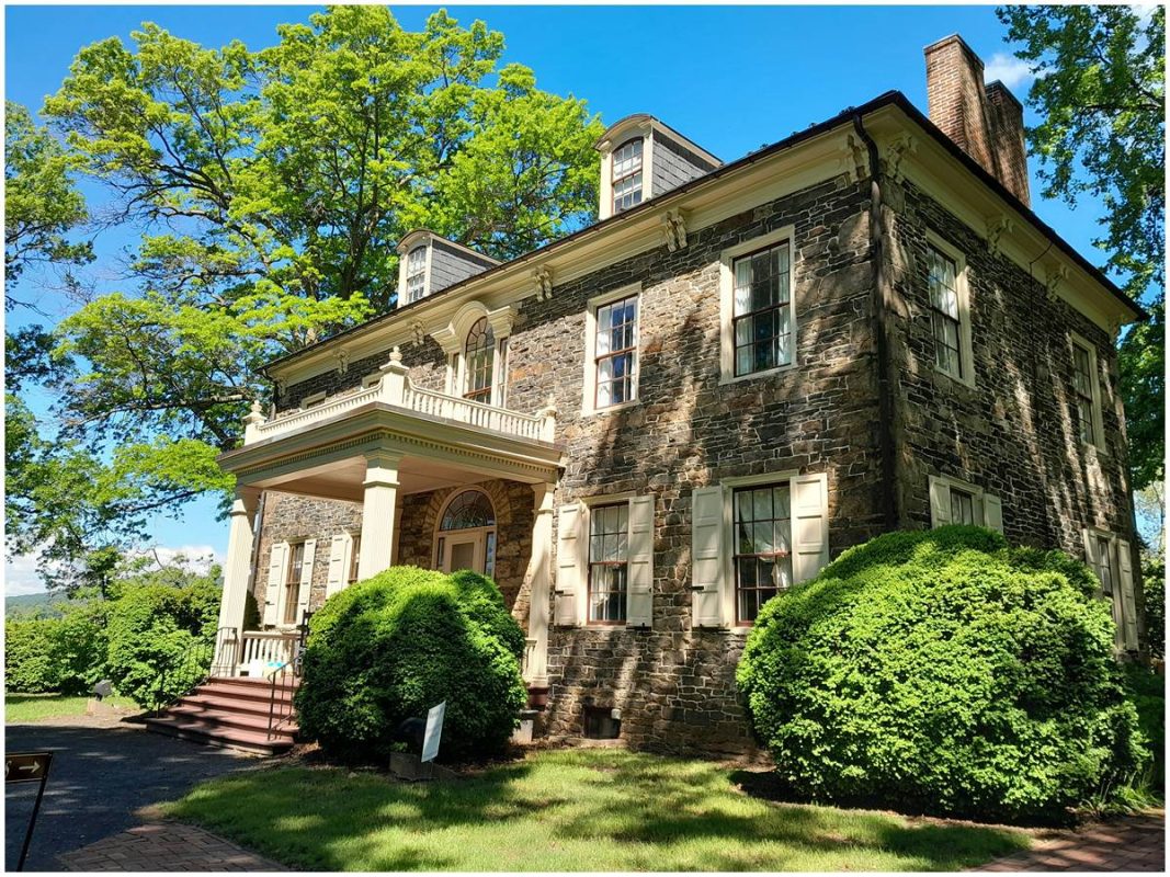 A stone manor with white columns and a covered porch, surrounded by trimmed bushes under a bright blue sky.